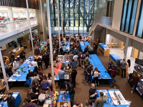 The image shows a large indoor event taking place in a spacious, modern venue with high ceilings and large windows. Numerous tables covered with blue tablecloths are set up, and people are gathered around them, engaging in various activities, possibly at a fair, exhibition, or conference. The scene is bustling with attendees of all ages, creating a lively atmosphere. The upper level of the venue has more seating and overlooks the event below.