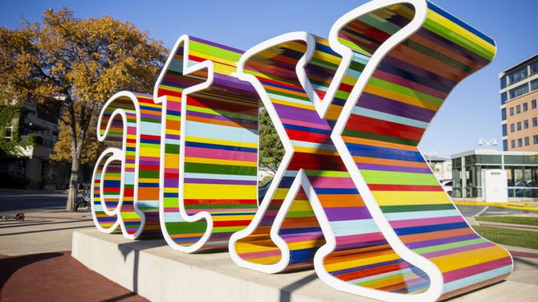 The image shows a colorful sculpture of the letters "atX" in a public outdoor space. The letters are decorated with horizontal stripes in various vibrant colors, creating a visually striking effect. The sculpture is mounted on a concrete base, with trees and buildings visible in the background under a clear blue sky. The setting suggests a lively urban environment, with the sculpture serving as an artistic and cultural landmark.