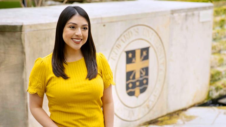 Victoria Garcia wears a yellow shirt and stands in front of the St. Edward's fountain and seal.