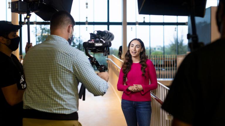 A woman with long, wavy hair wearing a bright pink sweater and jeans is standing and speaking in front of a camera crew indoors. She is facing the camera, with a man in a green checkered shirt holding the camera, and another crew member adjusting lighting equipment. The setting appears to be a spacious, well-lit area with large windows in the background, providing a view of the outdoors.