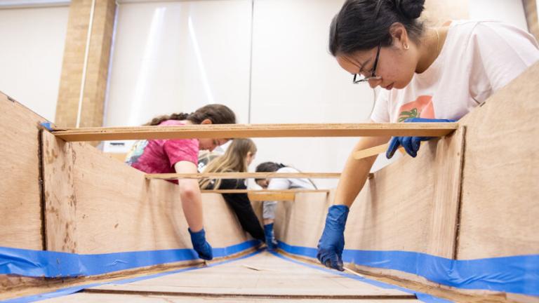 Several people are working on a wooden canoe structure indoors. They are focused on their tasks, applying material to the inside seams of the canoe. Each person is wearing gloves, with one woman in the foreground wearing a white t-shirt and glasses, carefully working on the edge of the canoe. The workspace is well-lit, with large windows providing natural light. Blue painter's tape is used to mark and protect certain areas of the canoe.