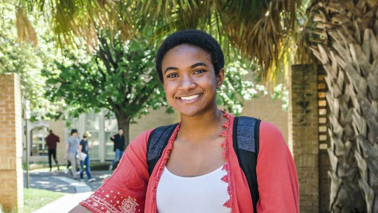 Jasmine Adgerson wears a white tank top and a red-pink blouse. Greenery and brick are in the background.