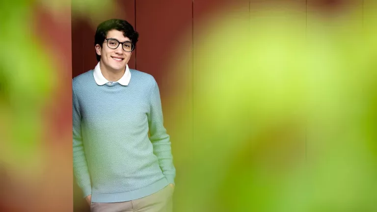 Cristobal Diaz wears a light blue sweater and glasses and leans on a red wall and is framed by blurry greenery.