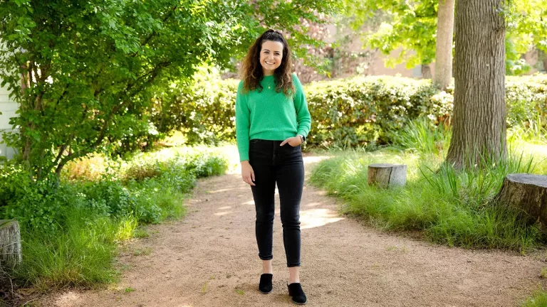 Jessica Guerriero wears a bright green sweater and black pants and stands in the Campus Ministry couryard, surrounded by trees and greenery.