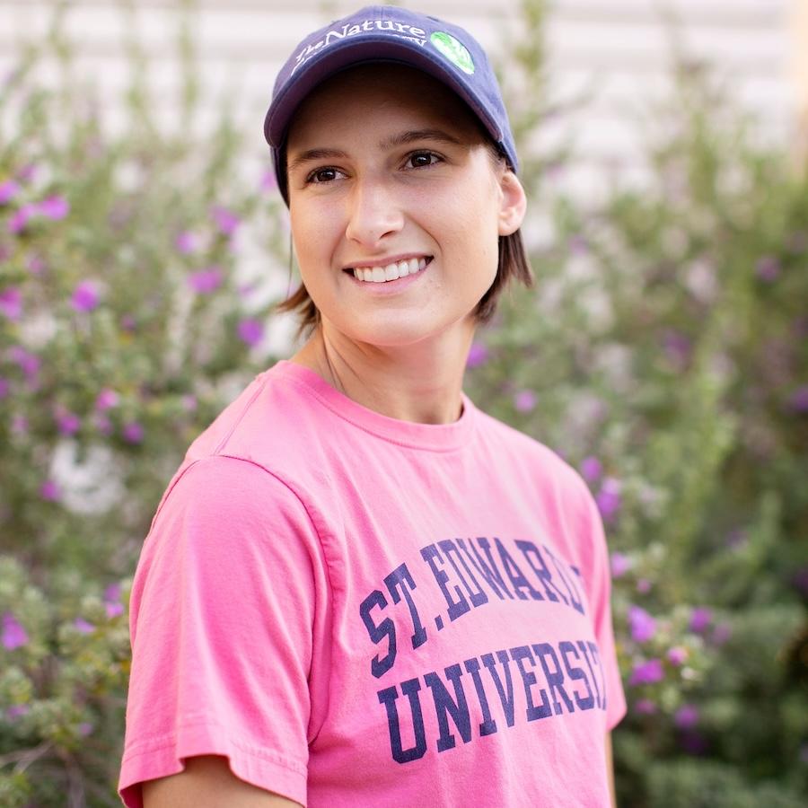 Stella Cunninghams wears a pink shirt that reads St. Edward's University in blue letters, as well as a blue cap that reads The Nature Conservancy. Blurred greenery and flowers are in the background.