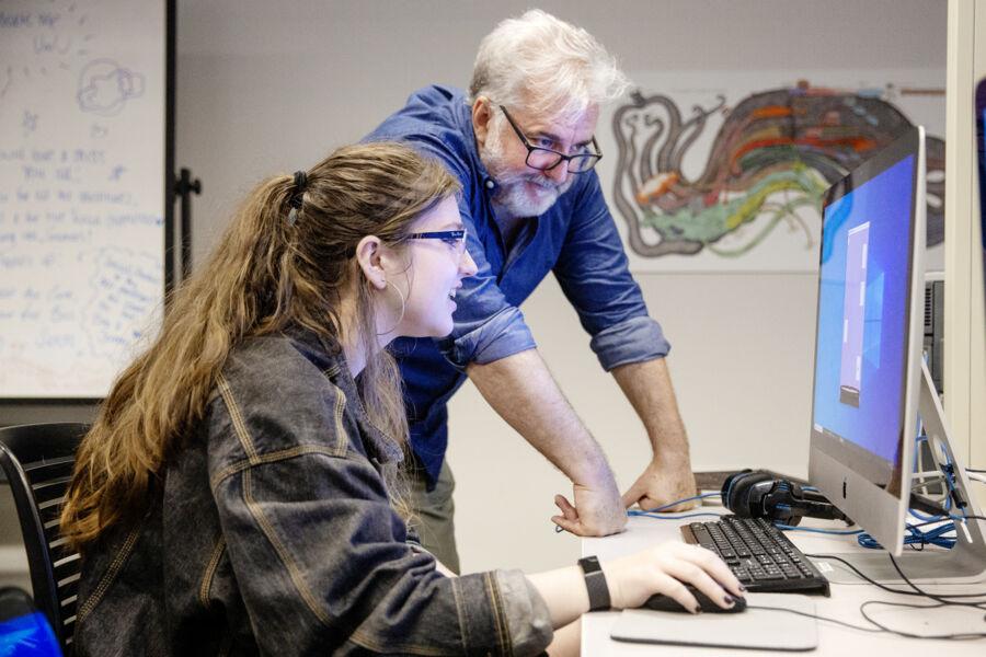 The image depicts two individuals in an office or classroom setting. The person on the left is seated, using a mouse, while the person on the right stands and points at something on the computer screen. In the background, a whiteboard displays writings and diagrams. The scene suggests collaboration, possibly teaching or project discussion.