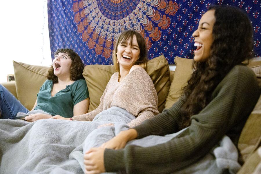 The image shows three young women sitting on a couch, laughing and enjoying each other's company. They are covered with blankets, indicating a cozy and relaxed setting. The background features a decorative tapestry with a colorful mandala pattern. One woman has curly hair and is wearing a green shirt, another has long, light brown hair and is wearing a beige sweater, and the third has long, dark hair and is wearing a dark green sweater. The atmosphere is joyful and warm, highlighting their close friendship.