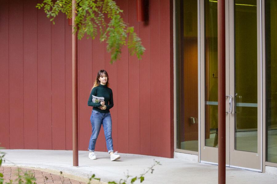 The image shows a young woman with long, light brown hair, wearing a dark green long-sleeve shirt, jeans, and white sneakers. She is walking outside along a curved walkway, holding several pieces of mail or papers. The building she is next to has a red exterior and large glass doors. The scene is framed with some greenery in the foreground, indicating an outdoor setting, likely on a campus. The woman appears focused as she walks.