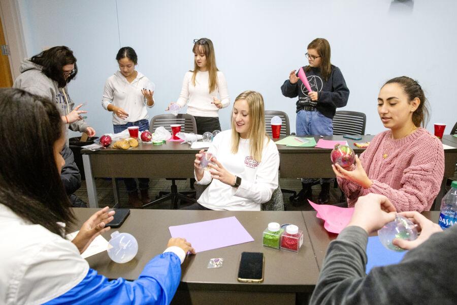 The image shows a group of people in a classroom-like setting, engaging in a craft activity. They are seated and standing around tables, decorating clear plastic ornaments with various materials such as glitter and colored paper. The participants, who include both men and women, appear focused and engaged in their tasks. The room has a casual atmosphere with supplies scattered on the tables, including scissors, glue, and additional craft materials.