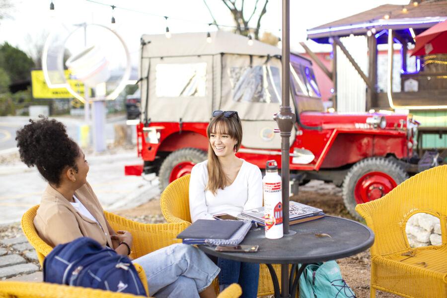 The image depicts two young women sitting at an outdoor café, engaged in a lively conversation. One woman has long, light brown hair and is wearing a white shirt, while the other has curly hair tied back and is wearing a tan jacket. They are seated at a round table with notebooks, a water bottle, and bags. In the background, a vintage red Jeep and some string lights add a charming, rustic feel to the setting. Both women are smiling, enjoying their time together in the casual, relaxed atmosphere.