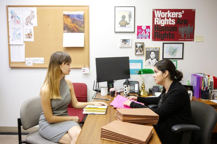 The image depicts two women in an office setting. One woman with long, light brown hair is wearing a sleeveless gray dress and is seated at a desk. She is engaged in conversation with another woman, who has dark hair tied back and is wearing a black top. The second woman is holding pink papers and has several brown folders stacked on the desk. The office is decorated with various posters and pictures on the wall. A corkboard with drawings and notes is also visible.