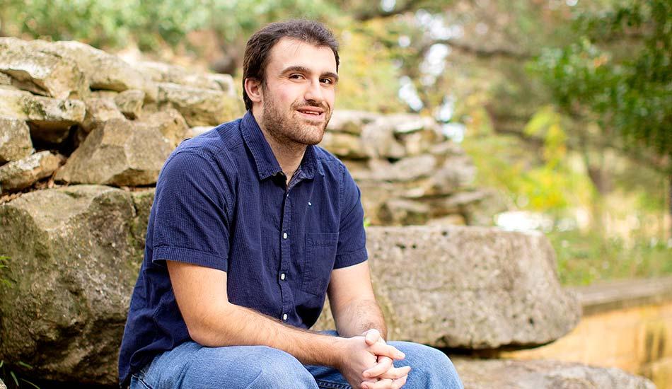 The image shows a man with short dark hair and a beard, wearing a dark blue short-sleeved shirt and blue jeans. He is sitting outdoors on a stone ledge with large rocks and greenery in the background. The man is smiling slightly and has his hands clasped together in front of him. The setting appears natural and serene, suggesting a calm and relaxed mood.