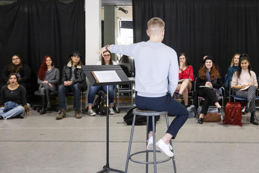 The image depicts a conductor leading a group of seated individuals. The conductor, facing away from the camera, uses their right hand to guide the group. Rows of audience members are visible. A music stand is in front of the conductor, suggesting a rehearsal or performance setting. The background features black curtains, indicating an indoor venue.