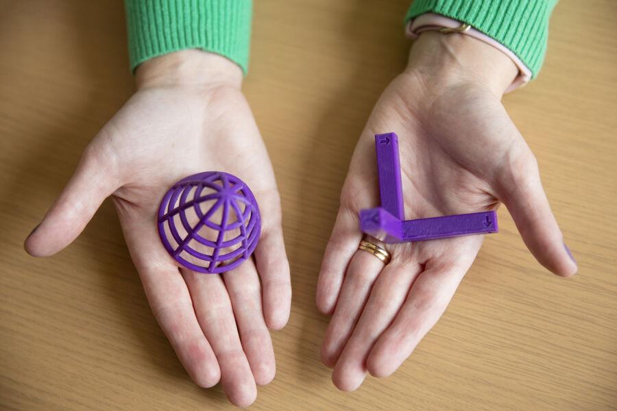 The image depicts two hands resting on a wooden surface. In the left hand, there's a purple spherical object with a lattice-like structure. The right hand holds a purple angular object resembling a cube corner with missing edges. These 3D-printed objects showcase geometric shapes and structures, possibly highlighting advancements in 3D printing technology or concepts in geometry.