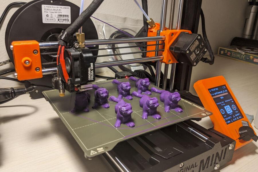 The image depicts a 3D printer with a print bed. On the bed, there are several small, identical purple figurines. The printer appears to be in the process of printing or has just finished printing these objects. Its orange and black color scheme contrasts with the background, which includes unrelated items like books or boxes. This image showcases the precision and capabilities of 3D printing technology.