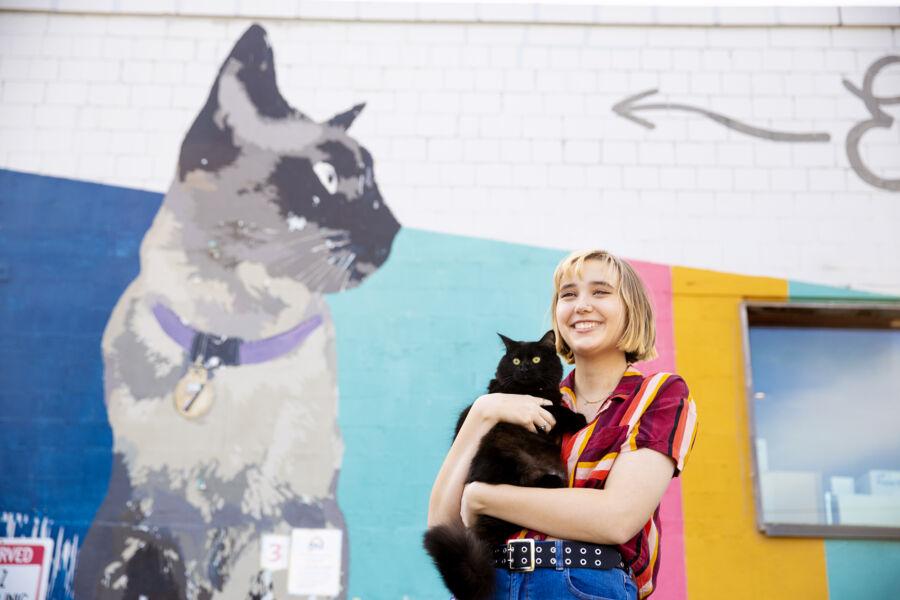 Ella Fotinos holds her cat and stands in front of a cat mural at Paz Vet.