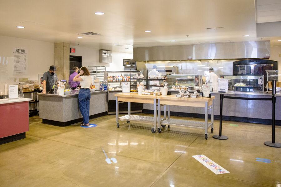 The image shows a spacious, modern cafeteria with various food stations. A few people are seen ordering and serving food, with one person in the foreground examining the options. The cafeteria has a clean and organized layout, featuring a selection of packaged foods, beverages, and freshly prepared meals. The floor is marked with social distancing indicators, and the atmosphere is calm and orderly. The setting suggests a focus on safety, efficiency, and a variety of dining choices.