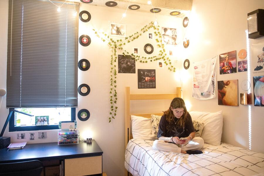 A student sitting on her bed in a brightly lit dorm with string lights and records on the wall