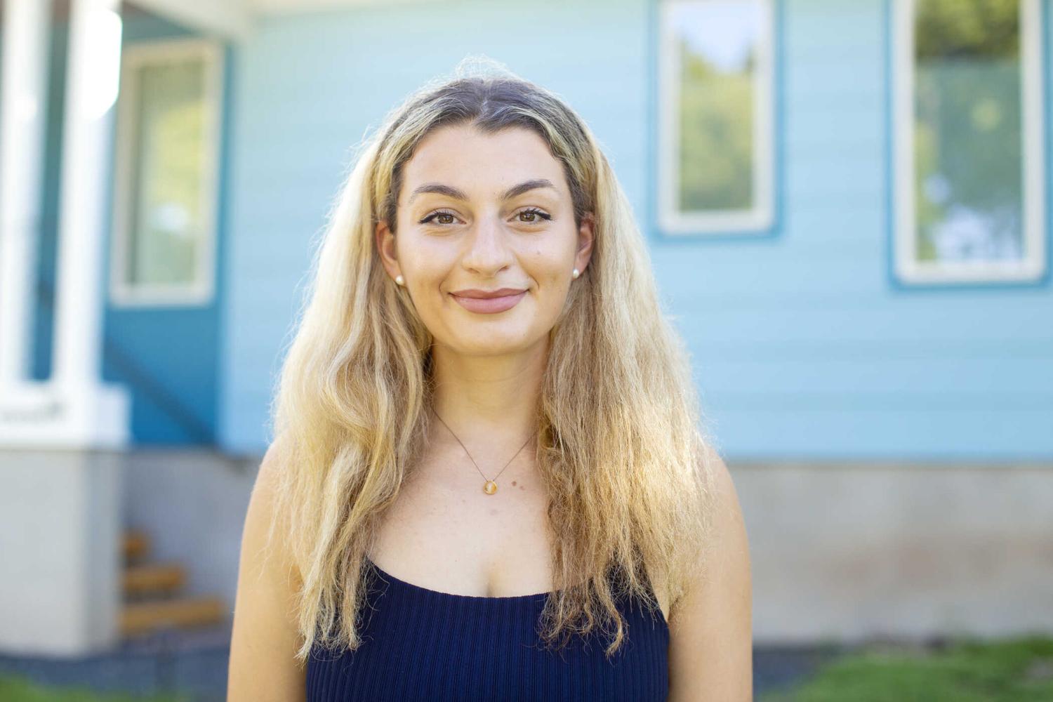 The image shows an individual standing in front of a house with blue siding and white trim. They are wearing a dark-colored top and a visible necklace. Behind them, there is greenery, suggesting a well-kept yard or garden area.