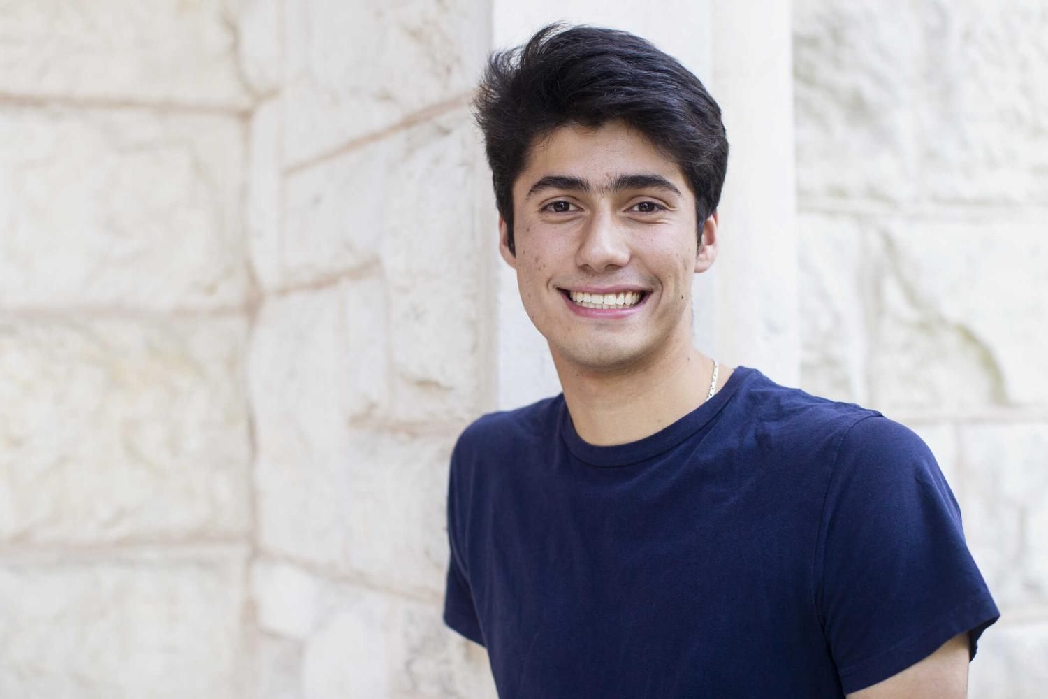 The image shows an individual wearing a dark blue t-shirt, standing in front of a light-colored wall with visible textures.