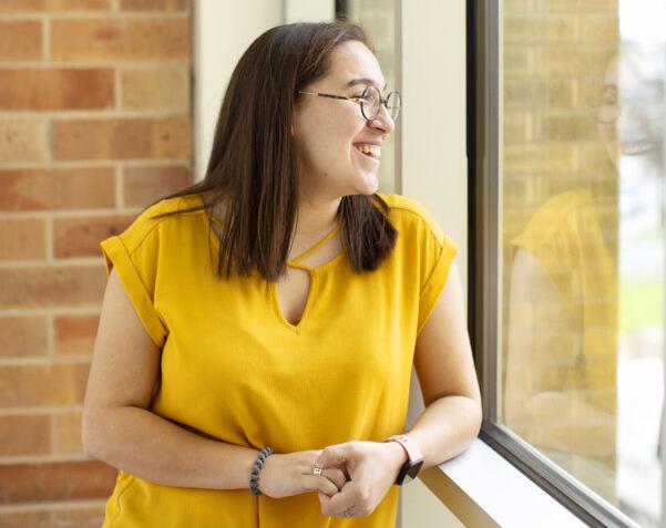 The image shows a woman with straight brown hair, wearing glasses and a yellow short-sleeved blouse. She is smiling and looking out of a large window. She is standing indoors, next to a brick wall, and her reflection is visible in the glass. She has a bracelet on her left wrist and is casually resting her right hand on the window sill. The overall mood is cheerful and relaxed.