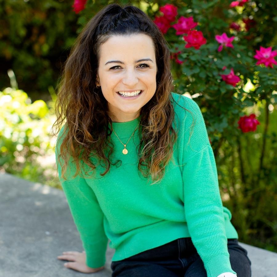 Jessie Guerriero wears a bright green sweater and black pants and sits in the Campus Ministry courtyard. Greenery and roses are in the background.