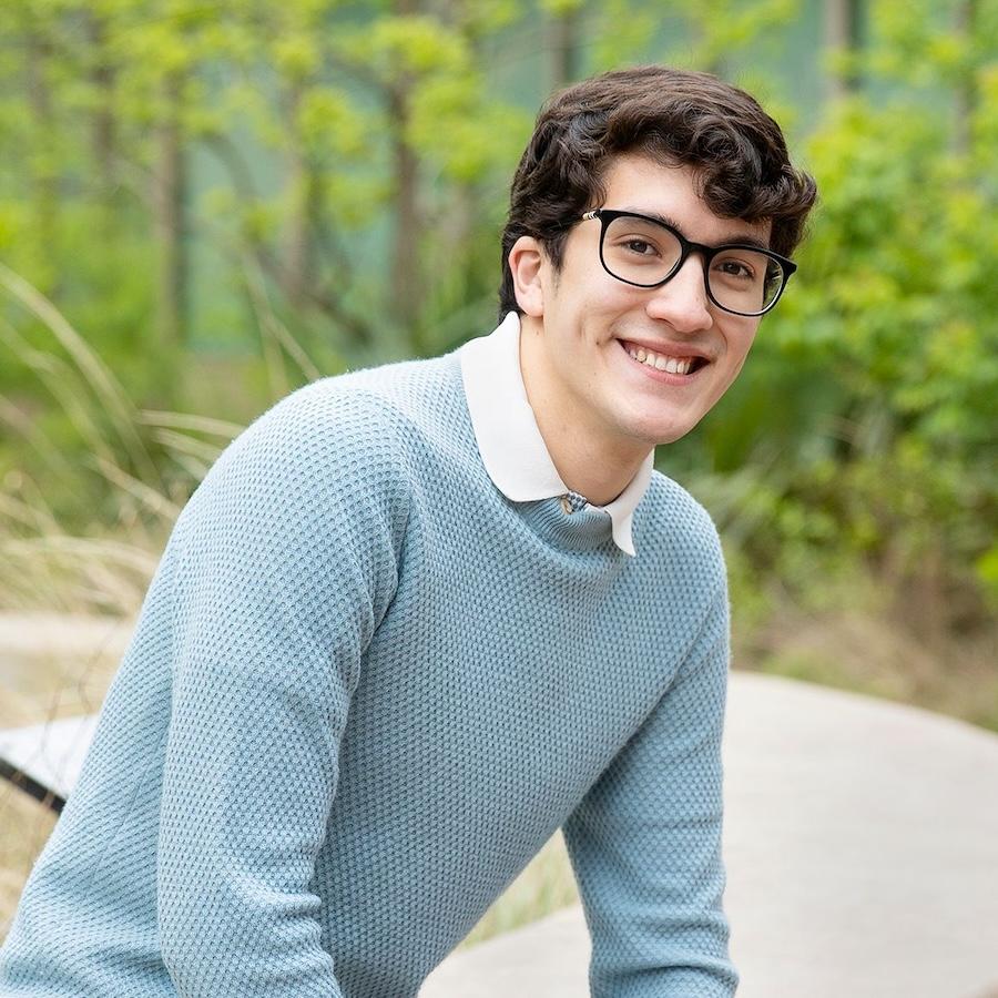 Cristobal wears a light blue textured sweater and glasses. Greenery is in the background.