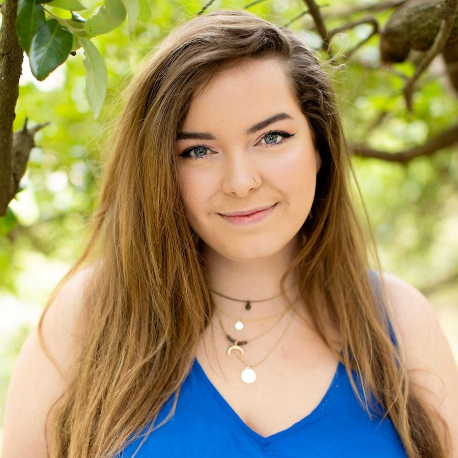 Jessica Riley wears a blue dress and multiple necklaces. Greenery is in the background.