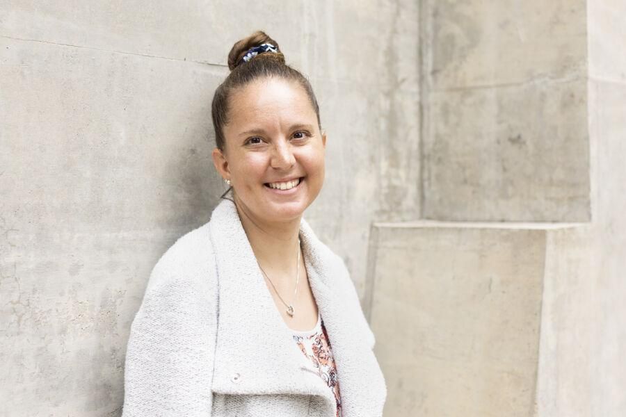 A woman in a white coat with her hair in a bun smiles at the camera in front of a concrete wall