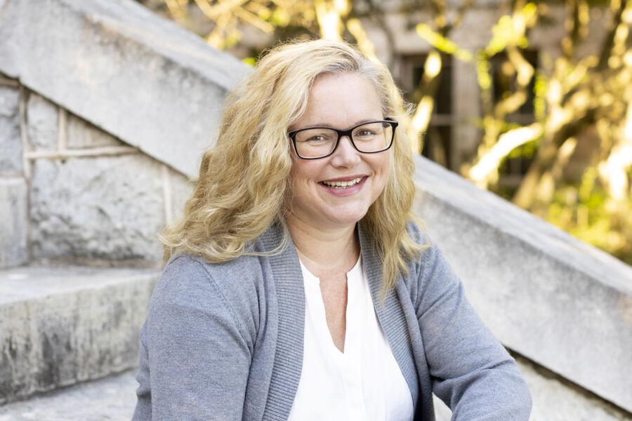 A blonge woman with glasses, sitting on an outdoor staircase, smiles at the camera