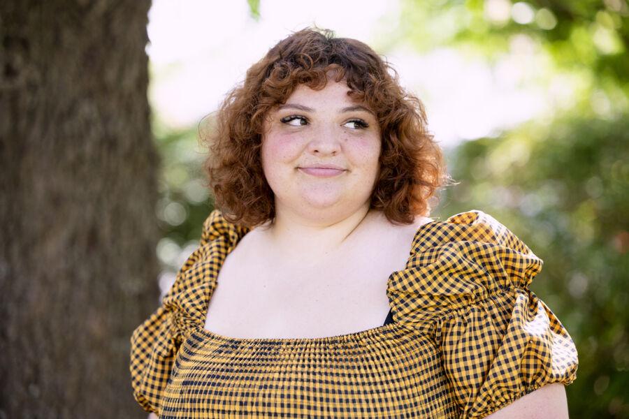 This image features a woman with curly brown hair and a cheerful expression, looking off to her right. She is wearing a yellow and black checkered off-the-shoulder dress with puffed sleeves. The background is a blurred outdoor setting with green foliage and a tree trunk. The lighting suggests a sunny day, enhancing the natural, relaxed atmosphere of the portrait.