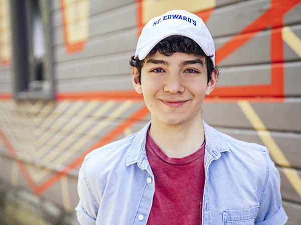 The image features a young man with short, dark curly hair wearing a white baseball cap with "St. Edward's" written on it. He is smiling and dressed in a light blue button-up shirt over a red T-shirt. The background is a mural with geometric patterns and a mix of gray, red, and yellow colors, painted on a wooden wall. The setting appears to be outdoors, creating a vibrant and casual atmosphere for the portrait.