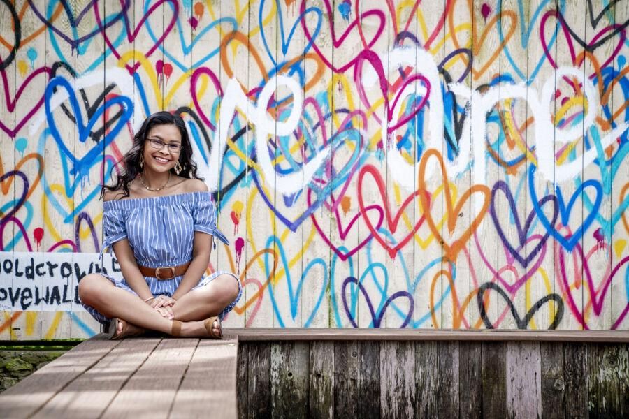 The image features a young woman with long, dark, wavy hair wearing glasses. She is smiling and dressed in an off-the-shoulder, blue and white striped top with short sleeves tied at the ends. She accessorizes with large, geometric earrings and a delicate necklace. The background is a vibrant mural with colorful heart shapes and abstract designs, adding an artistic and lively atmosphere to the image.