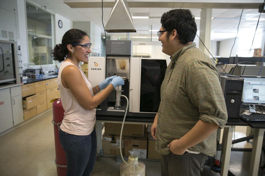 The image depicts a laboratory setting with two individuals. On the left, a person in a lab coat and gloves interacts with a machine, likely for chemical analysis. The person on the right observes. Various lab instruments and computers surround them, suggesting scientific research or testing. 