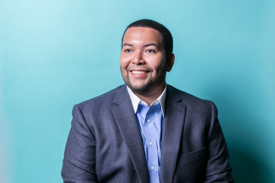 Dominick Namis wears a suit with a light blue backdrop behind him.