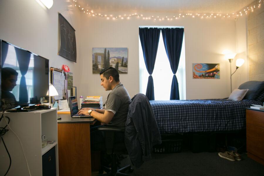 A student sitting at a desk at the end of a bed in a homely-lit dormroom