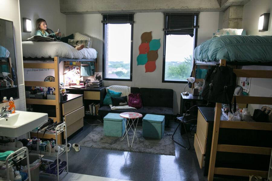 Two students sitting in stilted beds that have desks inderneath, with space in between them for a couch and chairs