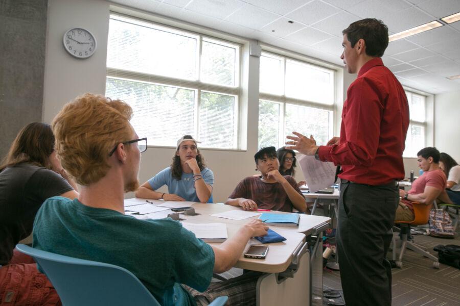 The image depicts a classroom setting. Several individuals, likely students, sit at tables with papers and notebooks. One person stands at the front, possibly the instructor. The clock on the wall shows approximately 10:10.