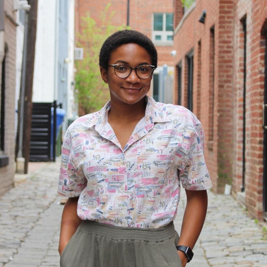 Jasmine Adgerson wears glasses, a colorful patterned blouse and pants and stands in an industrial alley.