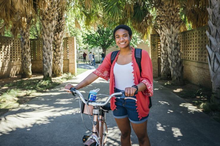 This image shows a young woman standing outdoors with a bicycle. She is smiling and wearing a red cardigan with decorative edges over a white top, paired with denim shorts. A backpack is strapped to her shoulders. The setting features a pathway lined with tall palm trees and brick walls, leading to a sunny, green area in the background. The scene has a relaxed and cheerful atmosphere, suggesting a pleasant day.