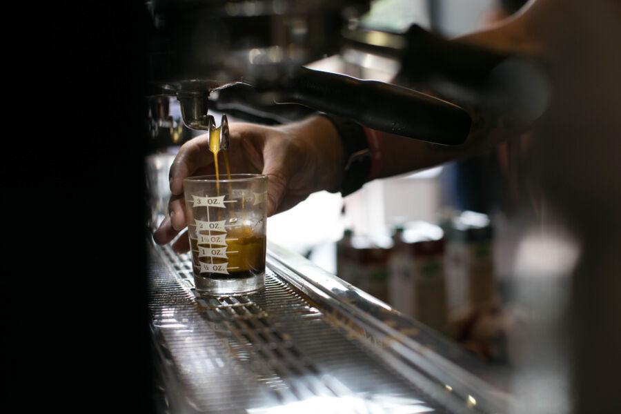 The image shows a close-up of a barista's hand pulling a shot of espresso using an espresso machine. The coffee is being dispensed into a small, clear measuring glass with markings indicating fluid ounces. The scene is set in a café environment, with the focus on the rich, dark espresso filling the glass, capturing the process of coffee preparation.