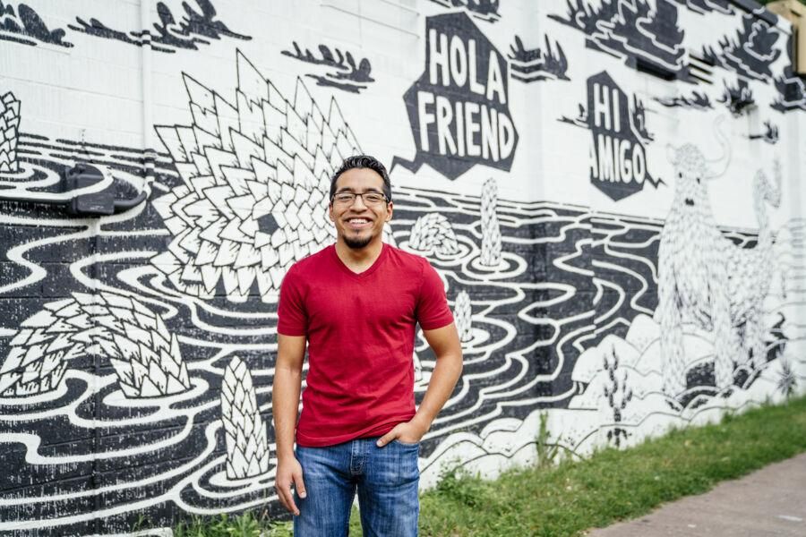 Joseph Ramirez stands in front of a black and white mural with creatures that say hola friend.