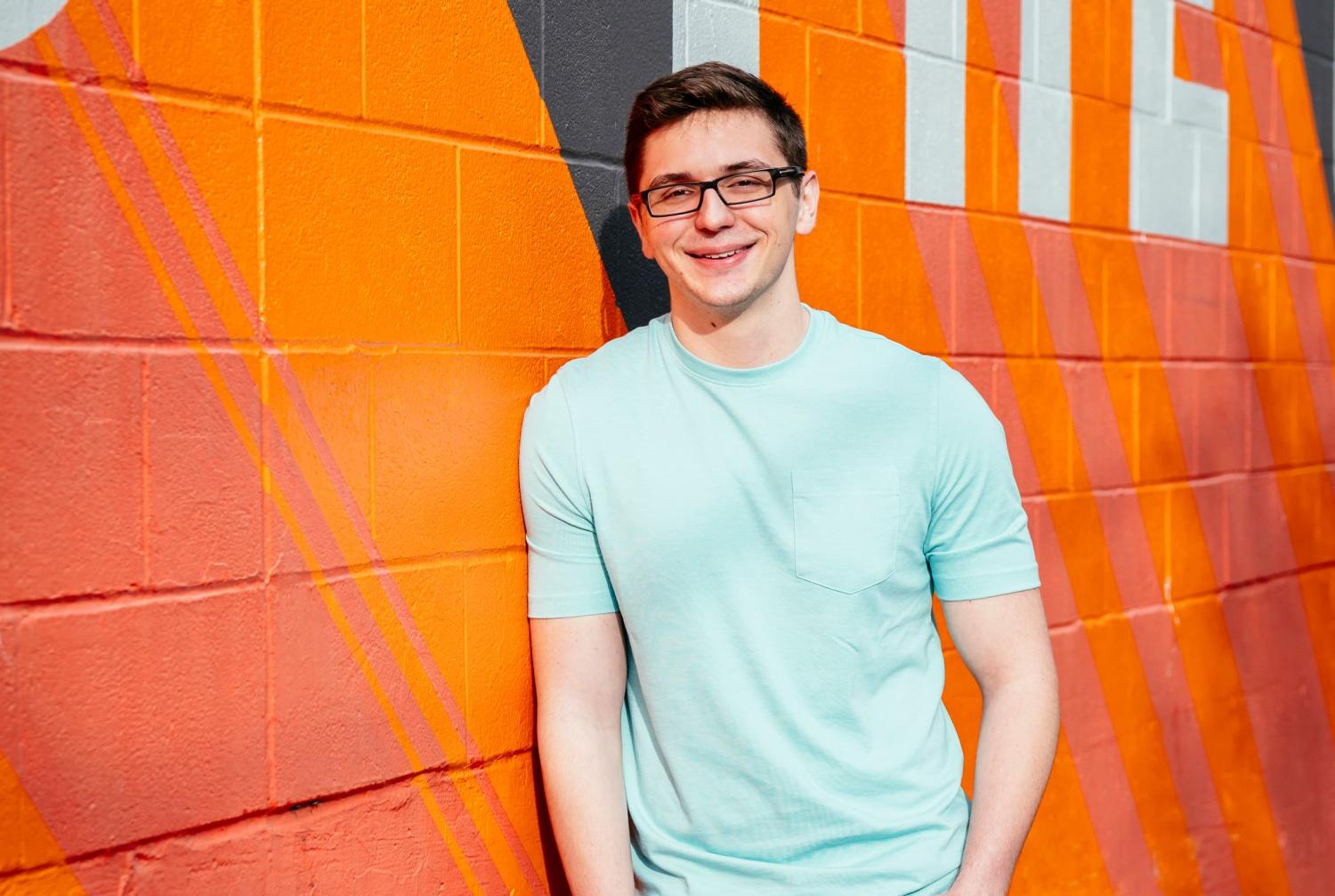 The image features a young man with short, dark hair and glasses, smiling at the camera. He is wearing a light blue T-shirt and standing against a colorful, vibrant mural wall painted with shades of orange, red, and black geometric patterns. The setting appears to be outdoors, with the bright mural adding an energetic and modern backdrop to the portrait.