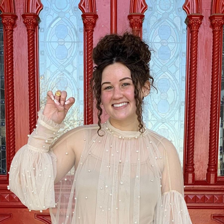 Lauren Louk wears a neutral, transluscent blouse with tank top underneath. They stand in front of the red doors and make a toppers up hand sign.