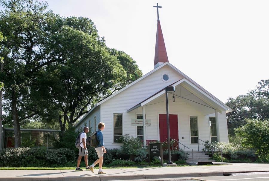 The image depicts a small white church with a red door and steeple. Trees surround the building, and two people walk past on the sidewalk. The church sign is partially visible. It's a serene scene, perhaps captured on a sunny day.