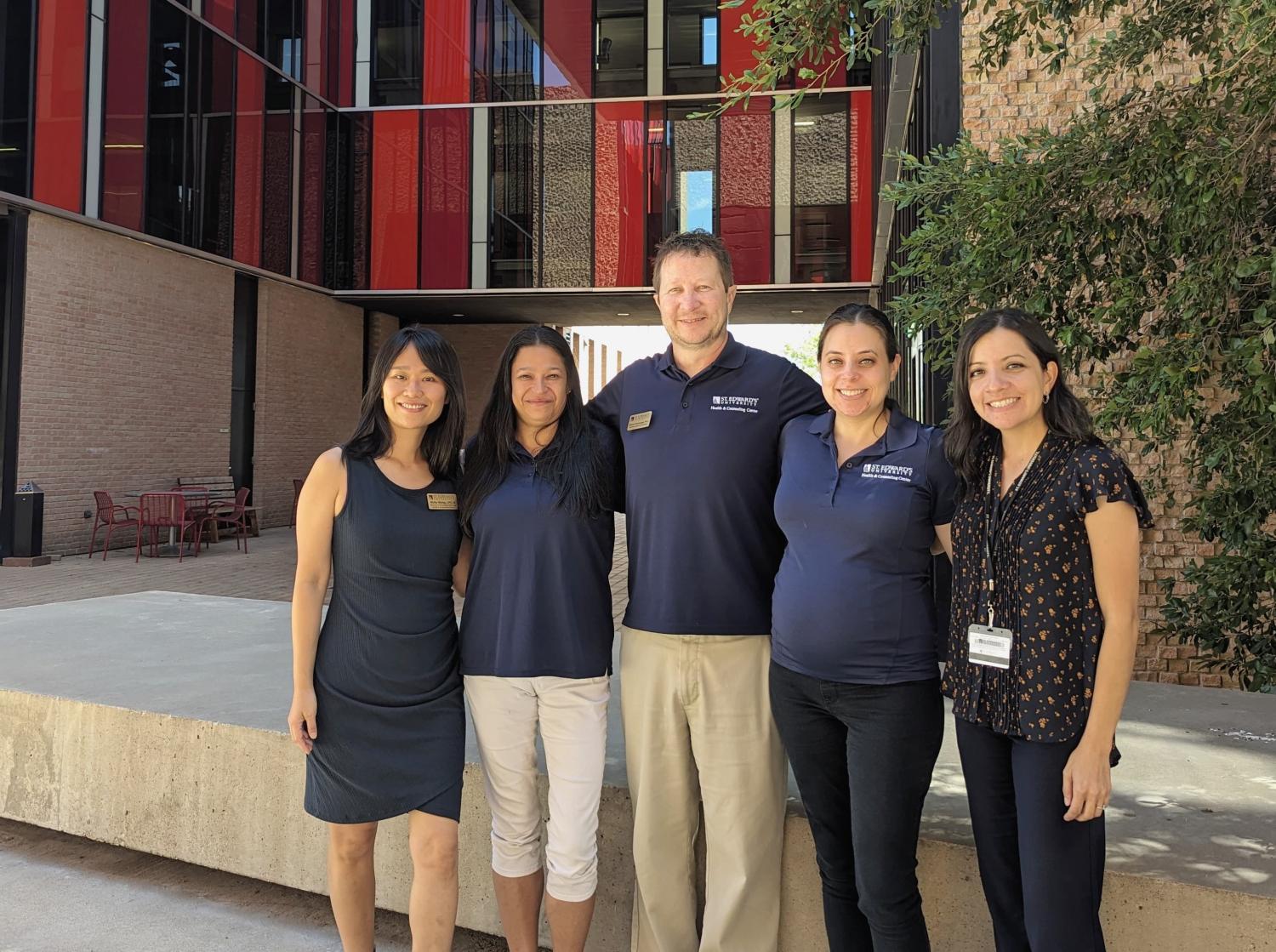 The image depicts five individuals standing in front of a modern building with red and grey accents. They are positioned on a concrete platform, wearing professional attire. The setting suggests a professional or academic environment, possibly related to an event or gathering at the building behind them.