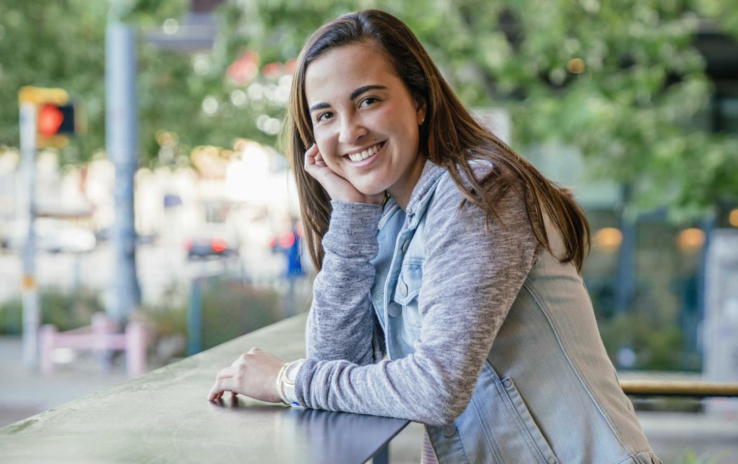 The image shows an individual sitting at a table. The person is wearing a light grey blazer over a darker grey or blue top. They have long brown hair that rests over their shoulders. The background suggests an outdoor setting, possibly an urban street scene, with trees and what appear to be traffic lights or street signs in the distance, slightly out of focus.