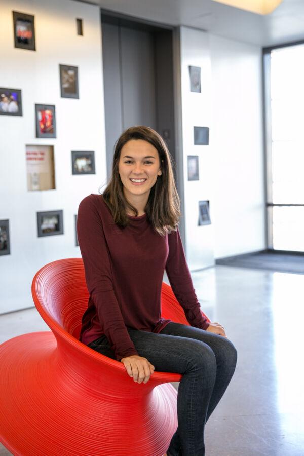 A woman in a burgundy sweater sits on an orange chair and smiles at the camera