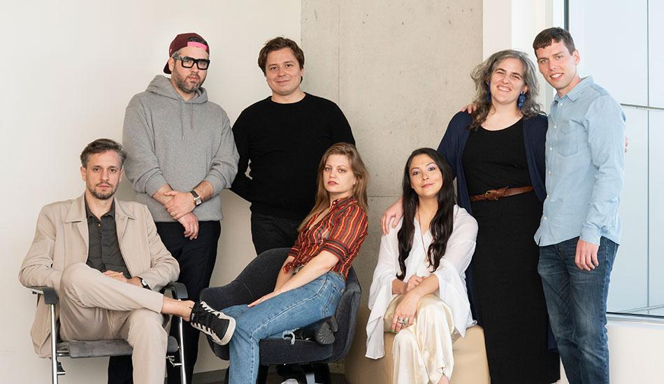 The image shows a group of seven people posing together indoors. Three are seated, and four are standing behind them. The seated individuals include a man in a light suit, a woman in a striped shirt and jeans, and a woman in a white blouse and light pants. The standing individuals include a man in a hoodie and cap, a man in a black shirt, a woman with gray hair in a dark dress, and a man in a blue shirt. They are positioned against a white wall and a concrete pillar, creating a casual and relaxed atmosphere