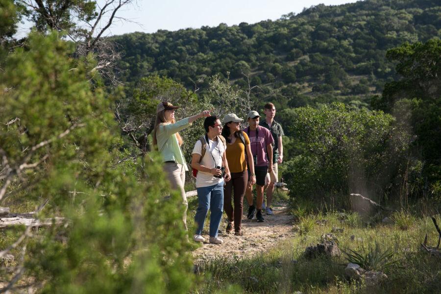 A group of 5 sfudtnes hiking through a park, looking over a hill